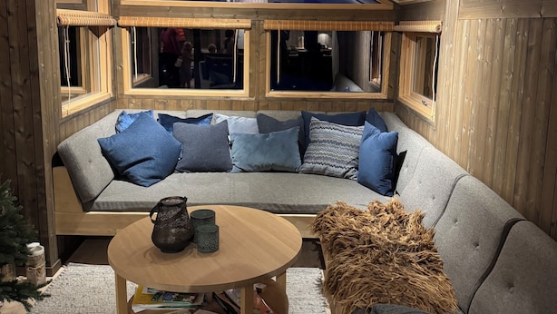 A cozy room with a wooden ceiling, a grey sofa with blue cushions, a round wooden table with a pitcher and books, and a woven pouf.