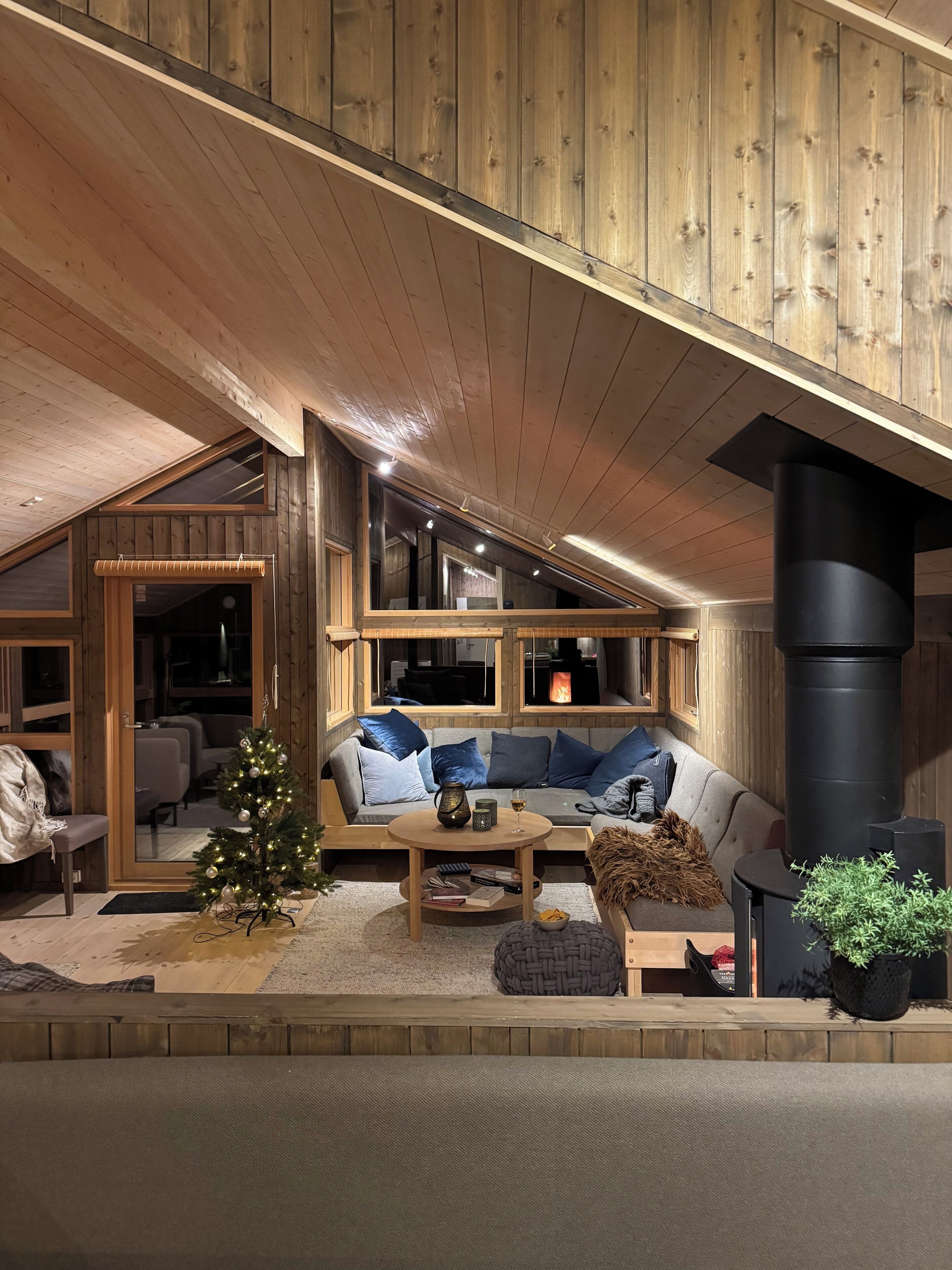 A cozy interior with a wooden ceiling, a fireplace, a sofa with cushions, and a small Christmas tree.