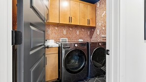 A compact laundry room with two front-loading washers and dryers, wooden cabinets above, and a white door on the left. - Reel & Ridge by Avantstay 2 Hot Tubs + Private Sauna + Movie Room + Fire Pit (Heber City)