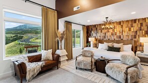 A modern bedroom with a large bed, two chairs, a nightstand, and a view of a winding road through a valley. - Reel & Ridge by Avantstay 2 Hot Tubs + Private Sauna + Movie Room + Fire Pit (Heber City)