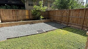 A residential backyard with a wooden fence, a grassy lawn, and a paved area with a drainage grate. - Cozy 1-bedroom apartment 4 mins from galleria mall Houston location (Houston)