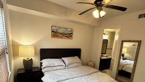 A bedroom with a bed, a ceiling fan, a nightstand, a lamp, a painting, a mirror, and a door. - Cozy 1-bedroom apartment 4 mins from galleria mall Houston location (Houston)