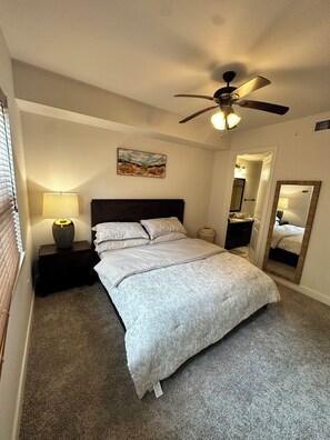 A bedroom with a bed, a ceiling fan, a nightstand, a lamp, a painting, a mirror, and a door. - Cozy 1-bedroom apartment 4 mins from galleria mall Houston location (Houston)