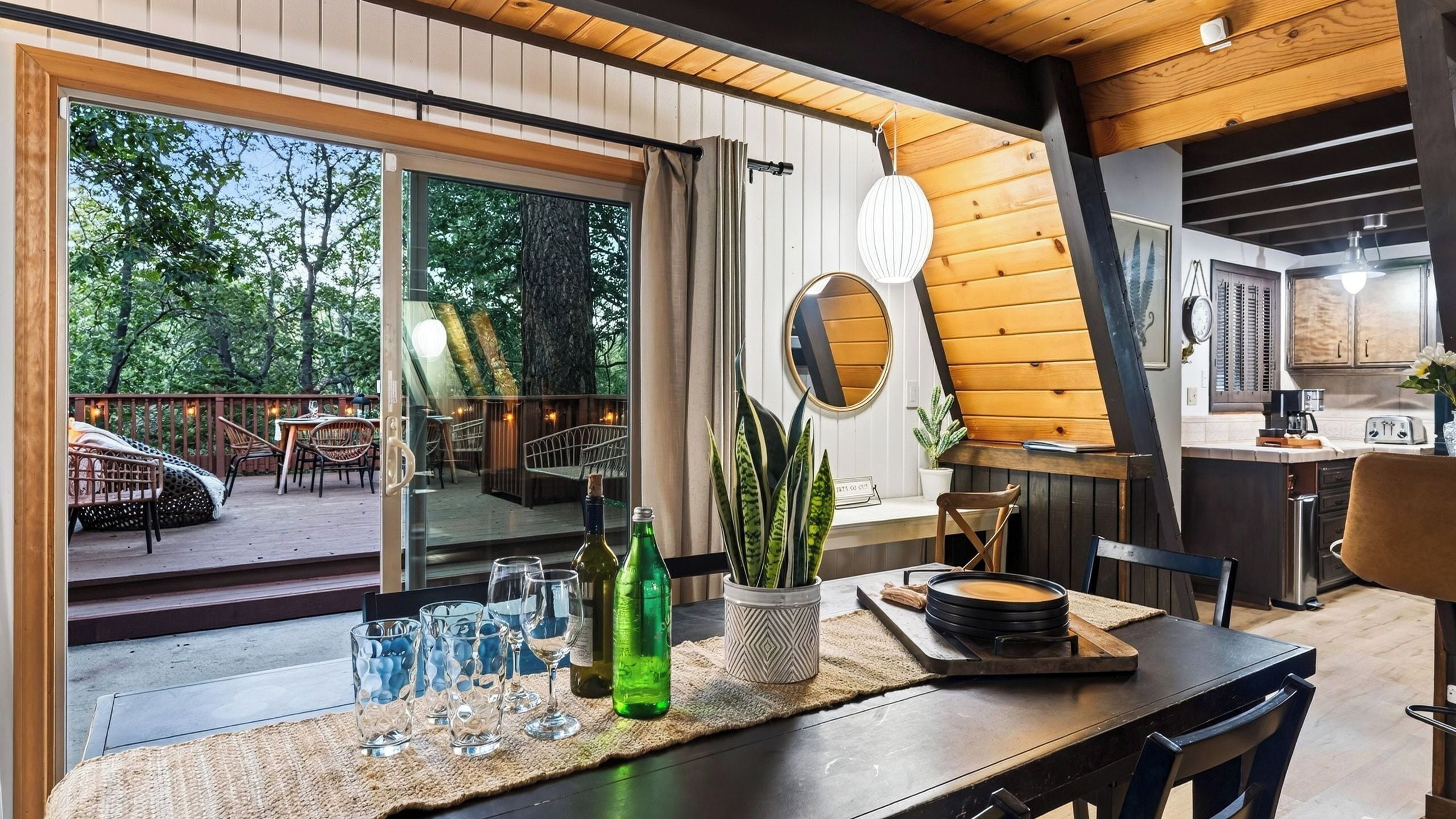 Un comedor moderno con una puerta de vidrio que da a una terraza exterior, un espejo redondo y una cocina con armarios de madera.