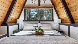 A bedroom with a sloped ceiling, a large window, a bed with a patterned bedspread, two bedside lamps, and a bedside plant. - A Big Bear View by Avantstay Classic A-frame Hot Tub Deck Fireplace Near Slopes & Marina (Big Bear Lake)