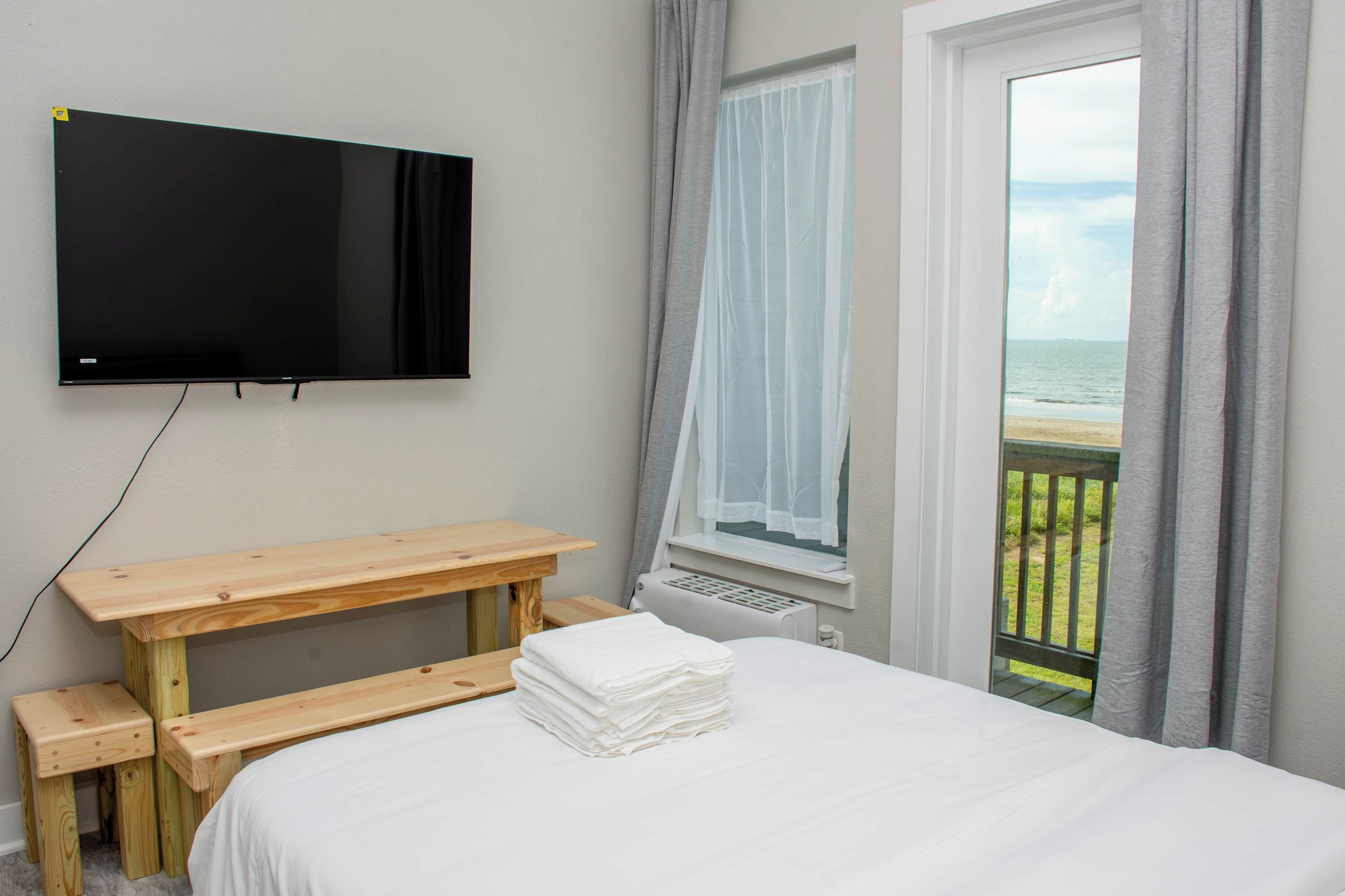 Une chambre à coucher avec un lit, une table en bois, un téléviseur fixé au mur et une fenêtre donnant sur la plage.