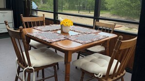 A dining area with a wooden table and chairs, a vase with yellow flowers, and a view of trees through large windows.