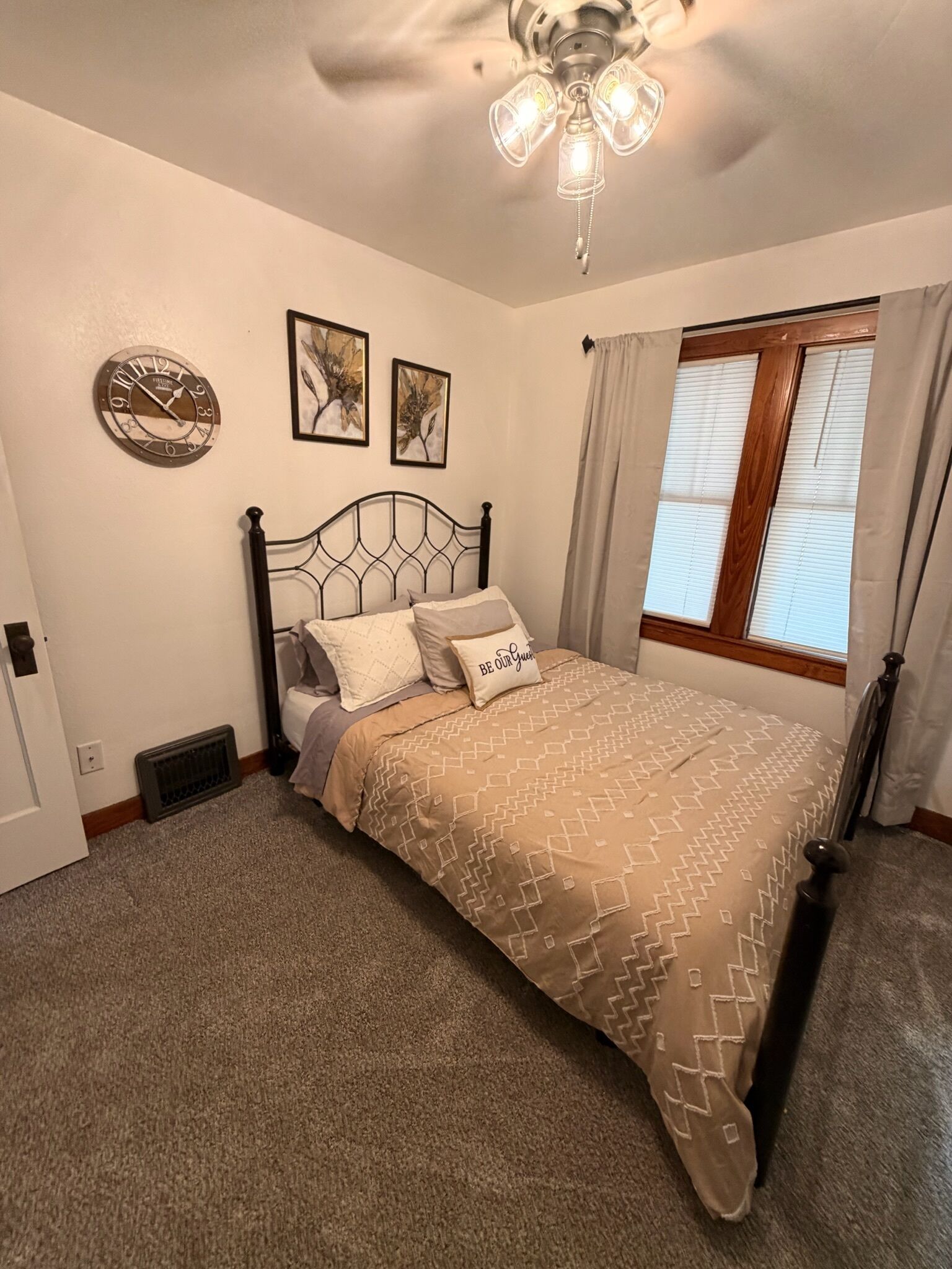 A bedroom with a bed, a wall clock, two framed floral pictures, a window with curtains, and a ceiling fan.
