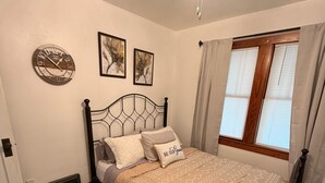 A bedroom with a bed, a wall clock, two framed floral pictures, a window with curtains, and a ceiling fan.