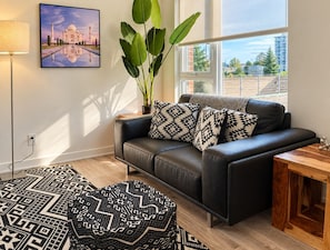 A modern living room with a black leather sofa, patterned cushions, a large potted plant, and a framed picture of the Taj Mahal. - Chic Urban 2BR Apt with Balcony and Skyline Views (Ottawa)