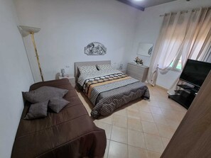 A bedroom with a bed, a brown sofa, a TV, a nightstand, and a lamp. - Charming Villa Rental in Mazara Del Vallo With Pool Access (Mazara del Vallo)