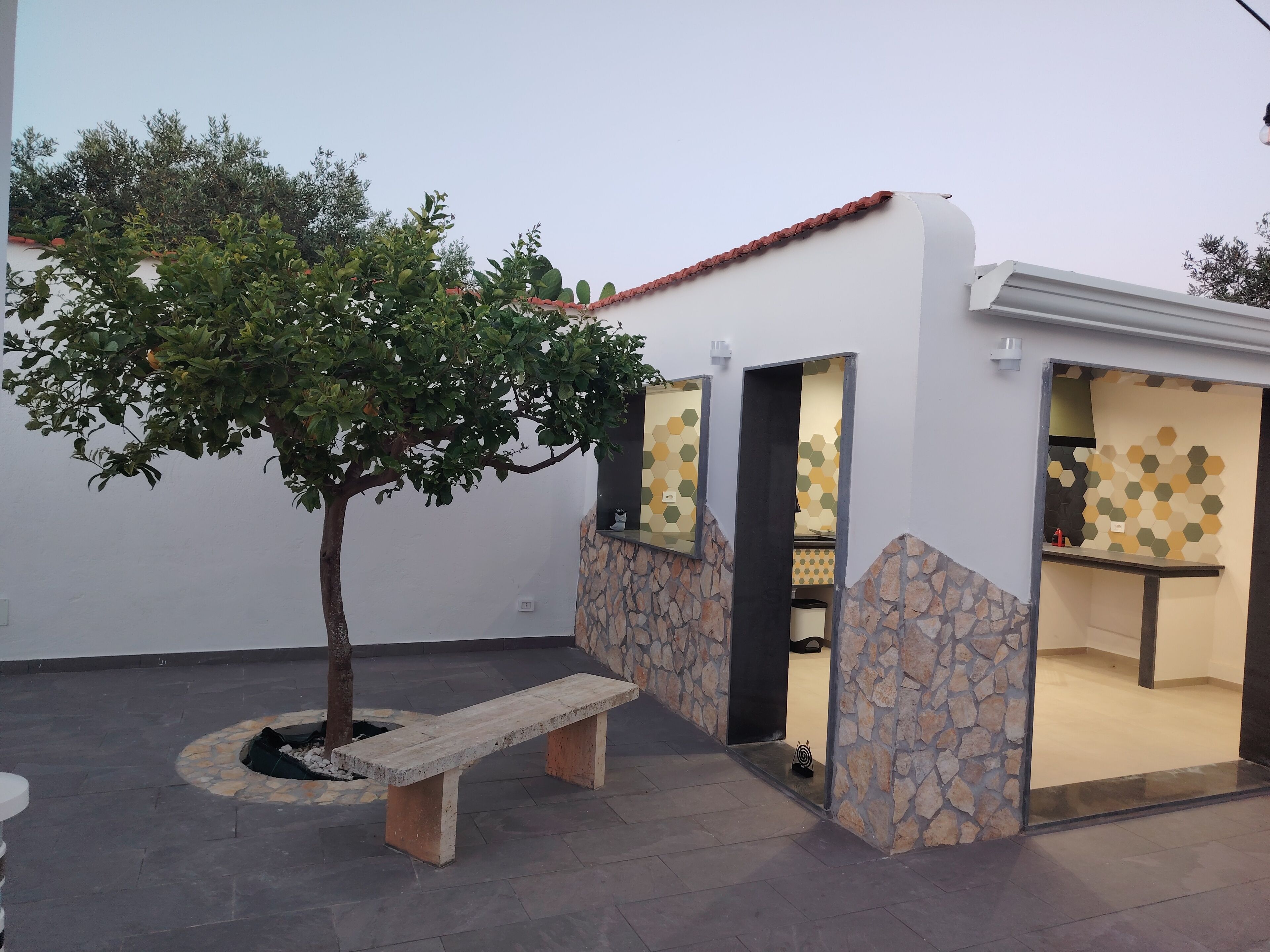 Ein kleiner Sitzbereich im Freien mit einer Bank, einem Baum und einem Gebäude mit einer Stein-Fassade und geometrischen Wandmustern.