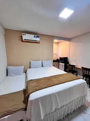 Une chambre d’hôtel avec deux lits simples, un lit double, un bureau avec une chaise, une kitchenette et un appareil de climatisation fixé au plafond.
