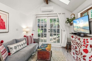 Ein Wohnzimmer mit grauem Sofa, einem farbenfrohen Couchtisch, einem Fernseher auf einer Kommode und einem großen Fenster mit Vorhängen.