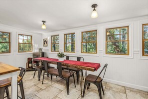 Ein Essbereich mit einem langen Tisch, Stühlen und einer roten Tischdecke. Es gibt Fenster mit Holzrahmen und Blick auf Bäume.