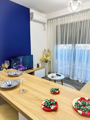 A modern living room with a TV, a dining table with plates and glasses, and a window with sheer curtains. - Hodo Apartments | Modern Cozy Apartment in Tirana 15 mins away from city Centre! (Tiranë)