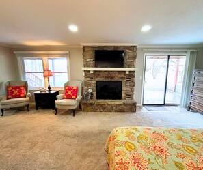 A bedroom with a stone fireplace, a television mounted above it, a bed with a floral bedspread, two armchairs with floral cushions, and a window with a view of the outside. - Stunning view of the North Golf Course from the back deck. (Cherokee Village)