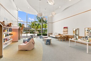 Ein geräumiger Spielbereich für Kinder mit großem Fenster, einer Couch, Tischen und einem Bücherregal.