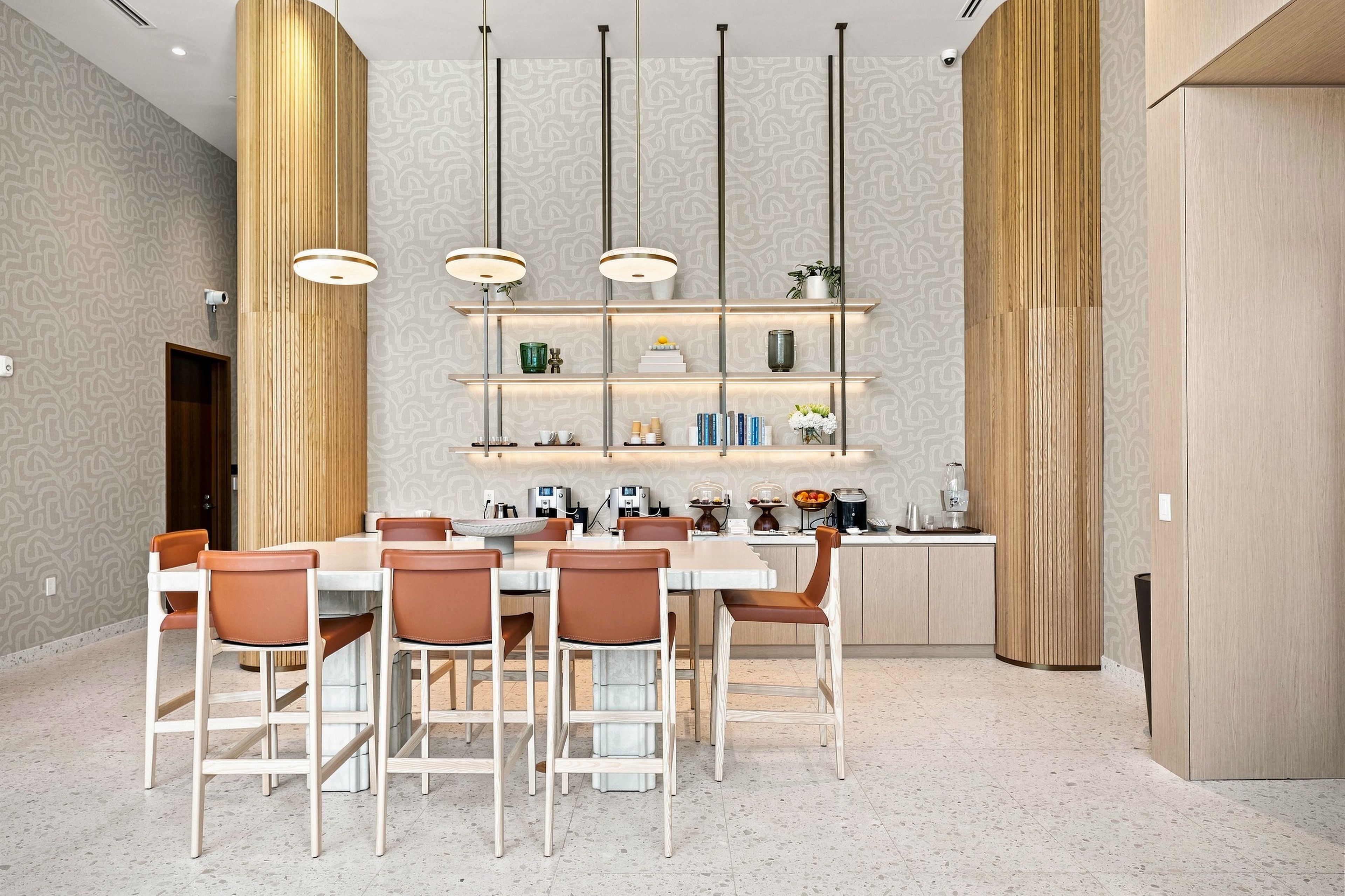 Uma zona de refeições moderna com uma mesa branca, cadeiras castanhas e uma ilha central com uma máquina de café e uma fruteira.