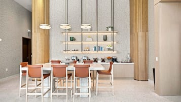 Uma zona de refeições moderna com uma mesa branca, cadeiras castanhas e uma ilha central com uma máquina de café e uma fruteira.