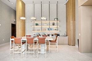Uma zona de refeições moderna com uma mesa branca, cadeiras castanhas e uma ilha central com uma máquina de café e uma fruteira.