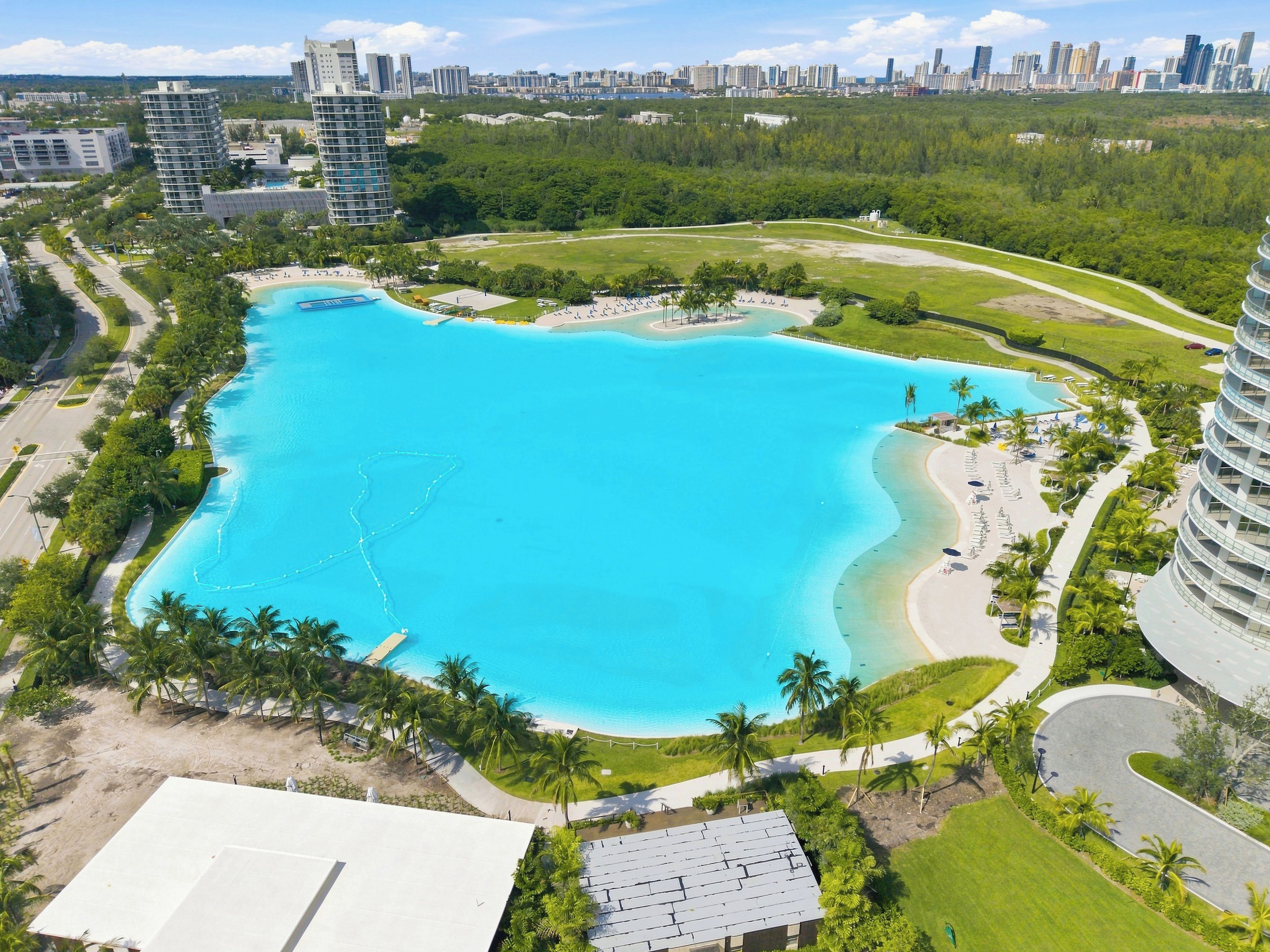 Uma piscina grande e límpida, azul, rodeada por palmeiras e com um horizonte urbano ao fundo.