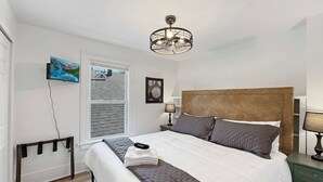 Een slaapkamer met een bed, nachtkastje, lamp, plafondventilator, raam en een aan de muur bevestigde televisie.
