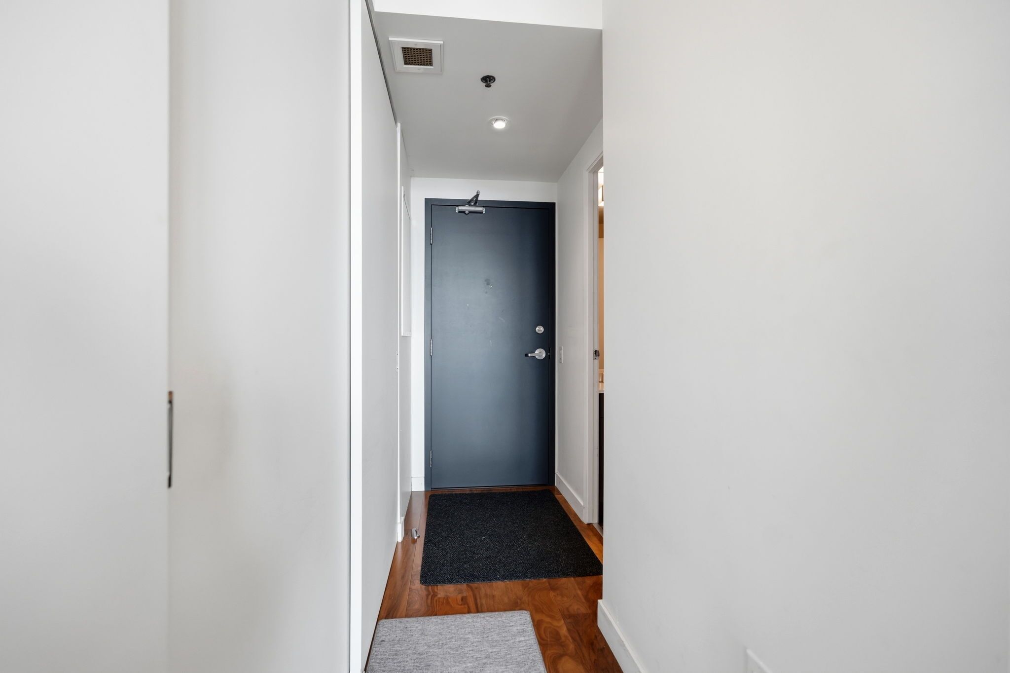 A narrow hallway with a dark blue door at the end, white walls, and a black mat at the entrance.
