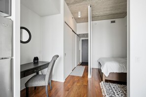 A compact room with a bed, a desk, and a chair. There is a door leading to another room. - Furnished Condo with Great Views | 1 Bed | 1 Bath | (Winnipeg)