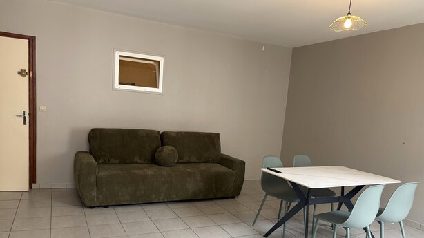 A room with a sofa, a dining table with chairs, a small window, and a light fixture. - Apartment in Downtown Montluel (Montluel)