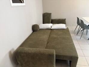 A room with a sofa bed, two pillows, a small round pillow, and a dining table with chairs. - Apartment in Downtown Montluel (Montluel)