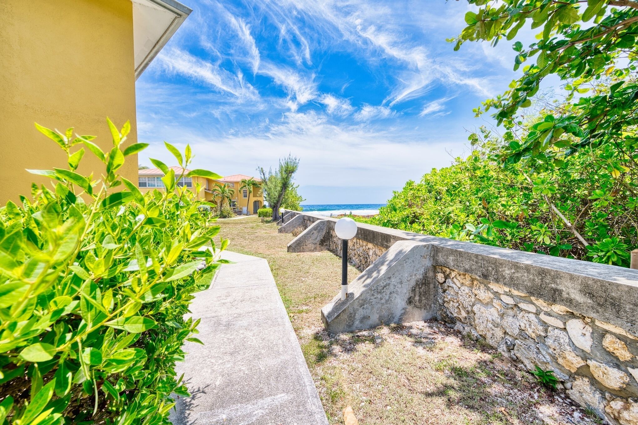 A pathway leading to a building with a view of the sea, surrounded by greenery and a clear sky.