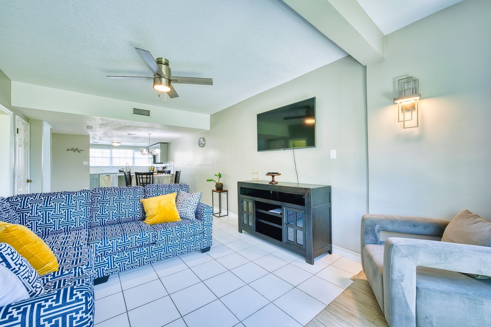 A living room with a patterned sofa, a grey armchair, a TV mounted on the wall, and a ceiling fan.