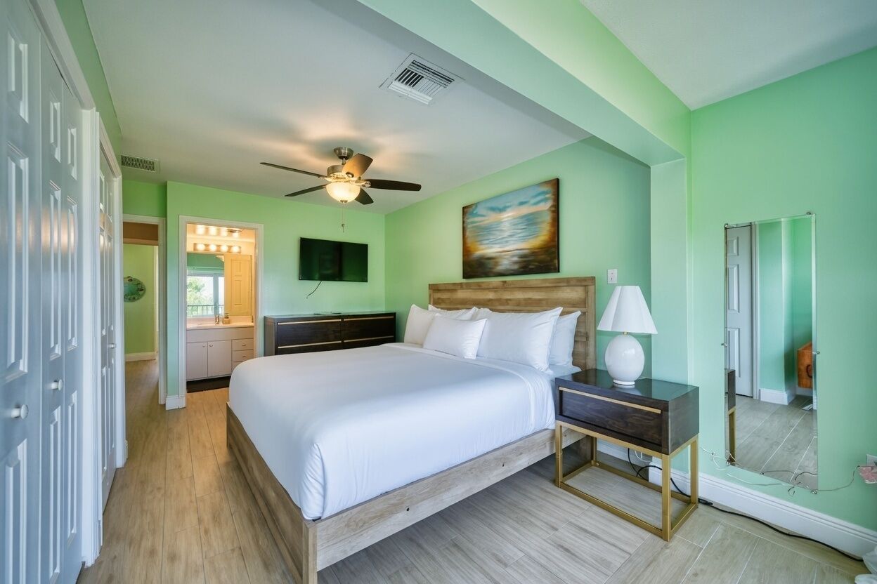 A bedroom with a bed, bedside table, lamp, ceiling fan, and a painting on the wall.