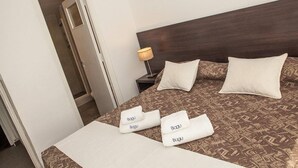 Habitación de hotel con cama, almohadas, mesita de noche con lámpara y toallas con la marca 'Bagu'.