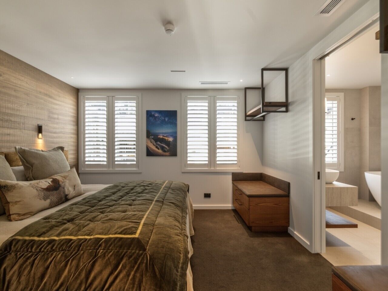 A bedroom with a bed, a nightstand, a wooden bench, a painting, and a bathroom with a bathtub.