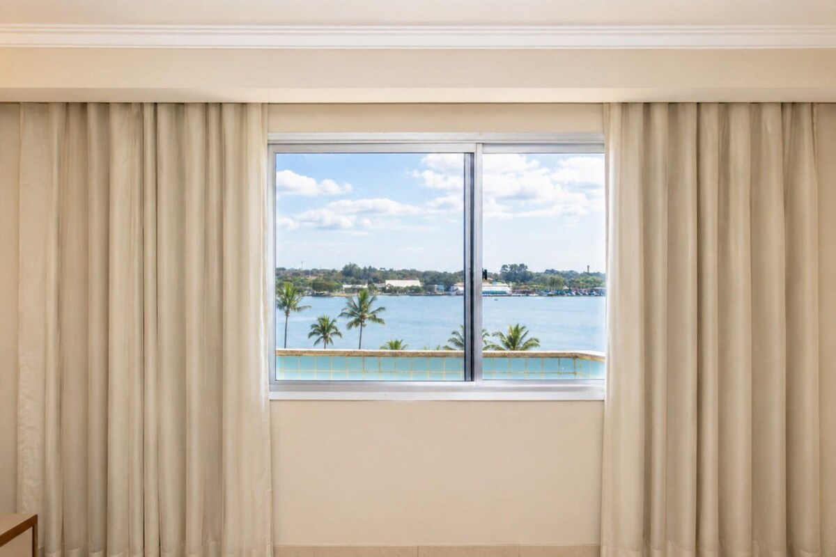 A room with a large window, curtains, and a view of a body of water with palm trees.