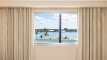 A room with a large window, curtains, and a view of a body of water with palm trees.
