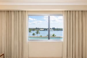 Una habitación con un ventanal, cortinas y vistas a un cuerpo de agua con palmeras.