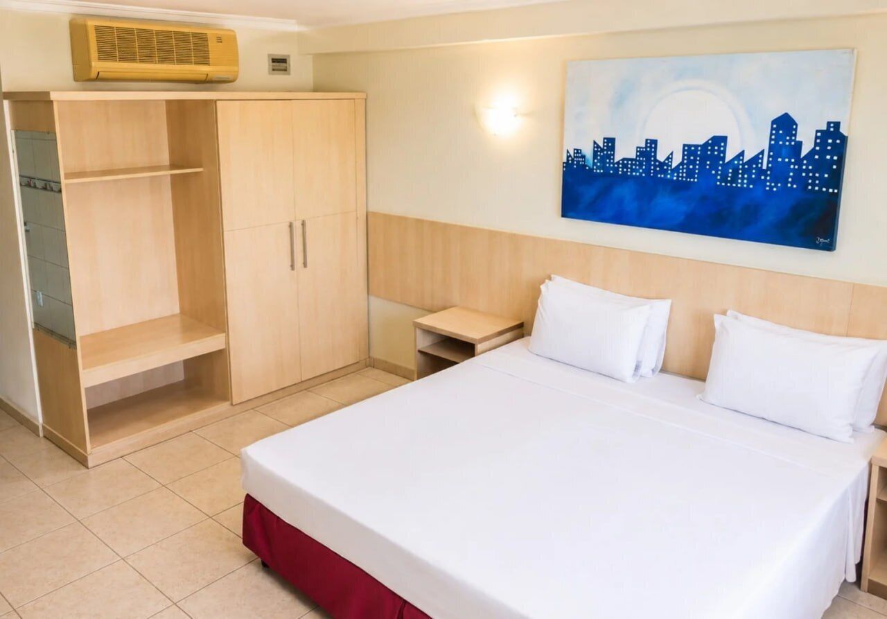 Habitación de hotel con cama, armario, mesita de noche y un cuadro de un paisaje urbano en la pared.