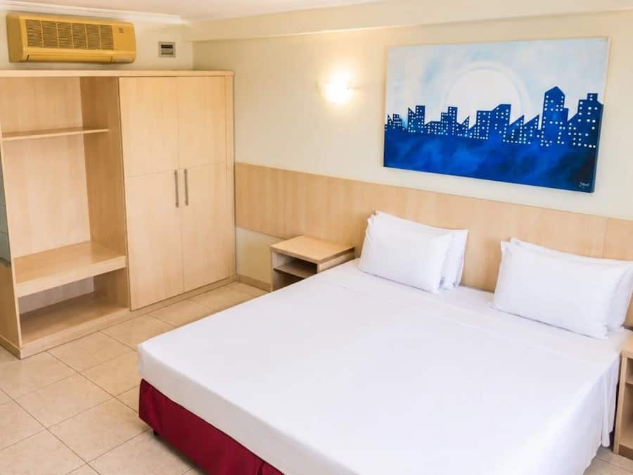 A hotel room with a bed, a wardrobe, a nightstand, and a cityscape painting on the wall.