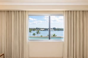 A room with a large window, curtains, and a view of a body of water with palm trees.