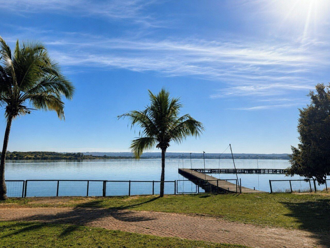 Un parque frente al agua con un paseo pavimentado, palmeras y un muelle que se adentra en una masa de agua tranquila.