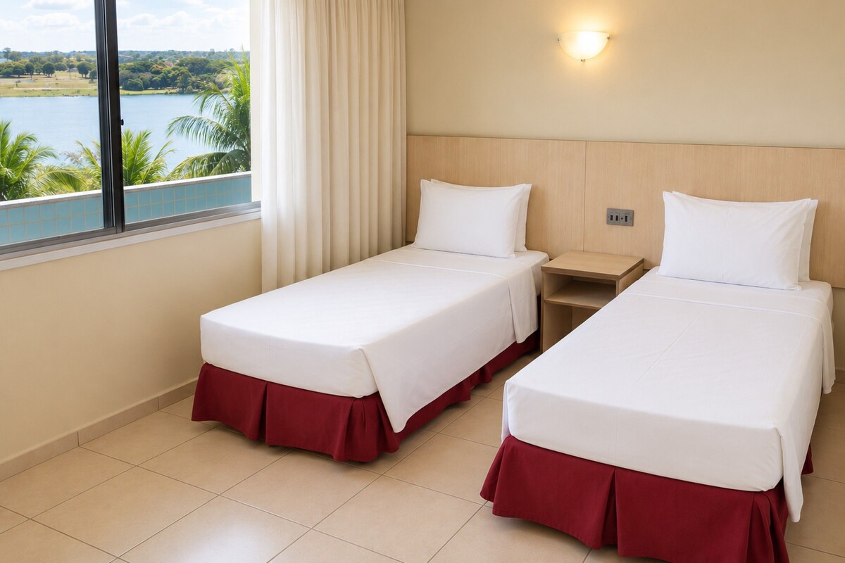 A hotel room with two single beds, a nightstand, a window with a view of a lake, and a wall-mounted light fixture.