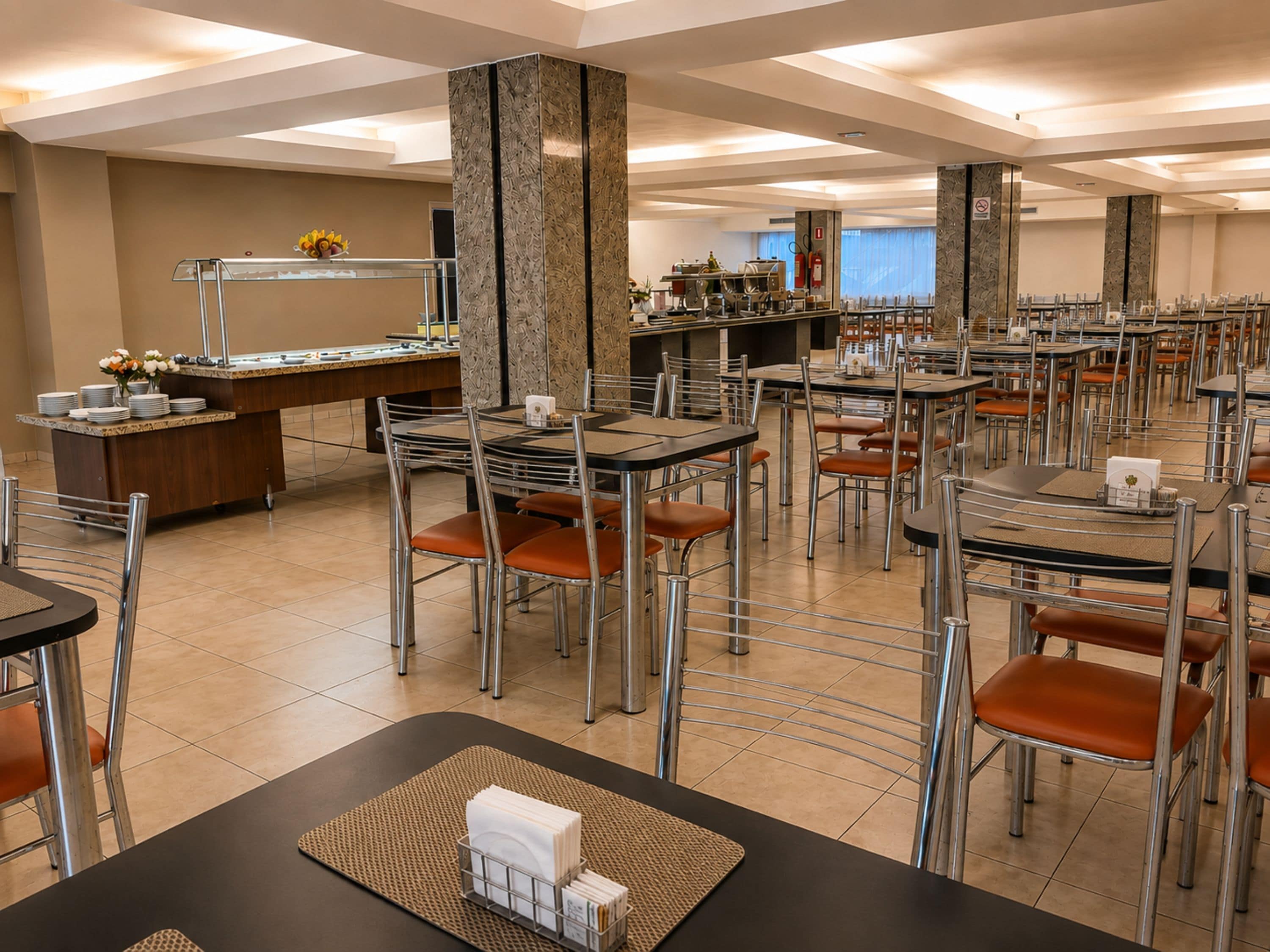 Un'area ristorante di un hotel con diversi tavoli e sedie, un'area buffet e una cucina sullo sfondo.