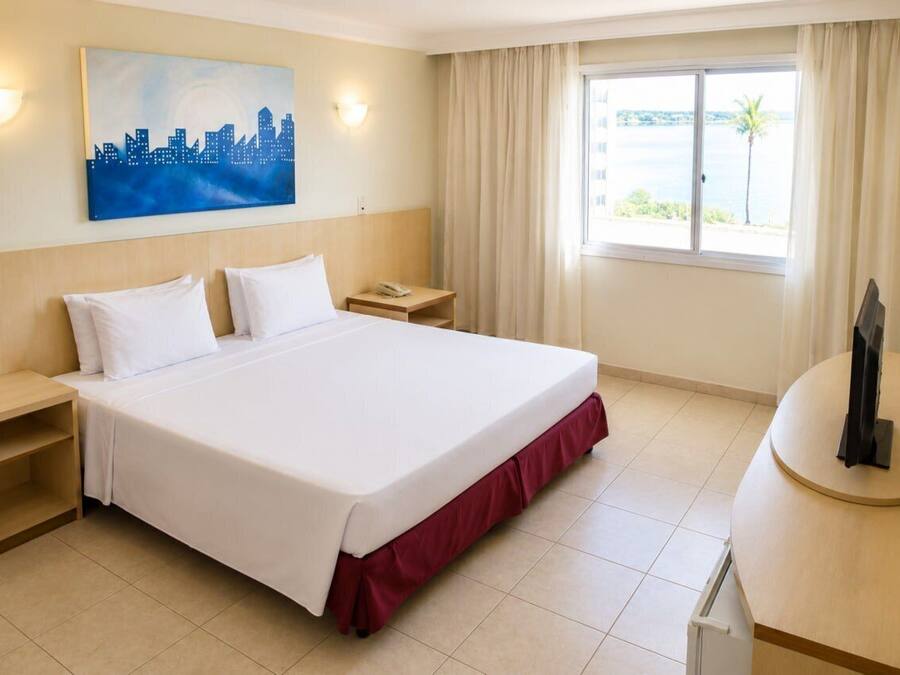 Quarto de hotel com cama, mesas de cabeceira, escrivaninha, cadeira, quadro e janela com vista.
