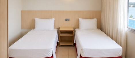 Habitación de hotel con dos camas individuales, una mesita de noche, un enchufe y una ventana con cortinas.