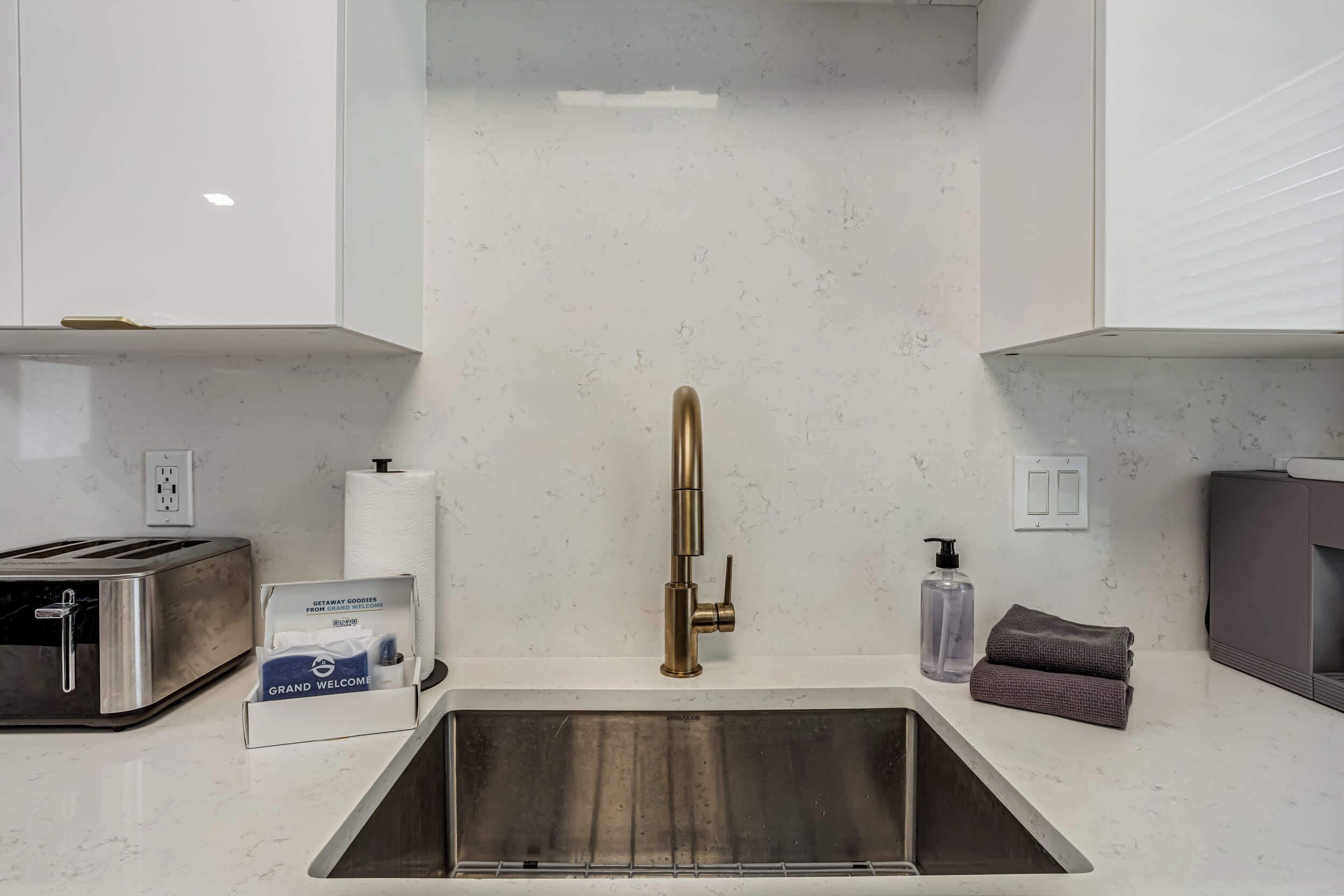 Una cocina con un lavamanos de acero inoxidable, un tostador, dispensador de toallas de papel y una botella de jabón para manos.