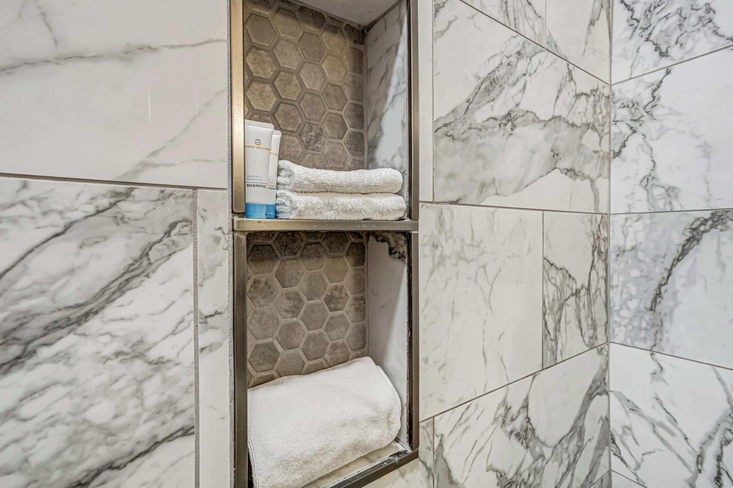 A bathroom with marble tiles, a shower area, and a shelf with toiletries.