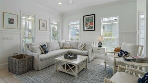 A spacious living room with a white sofa, a coffee table, and a chair. There are framed pictures on the walls, a ceiling fan, and a window with blinds. - Blue Mountain View | Private Pool, Rooftop Deck & Bikes (Santa Rosa Beach)